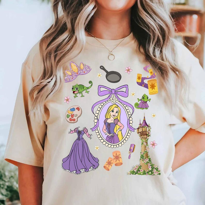 May include: Beige t-shirt featuring a collection of cartoon illustrations. The design includes a princess portrait, a tower, a dress, a frying pan, and other related elements in purple, yellow, and green.