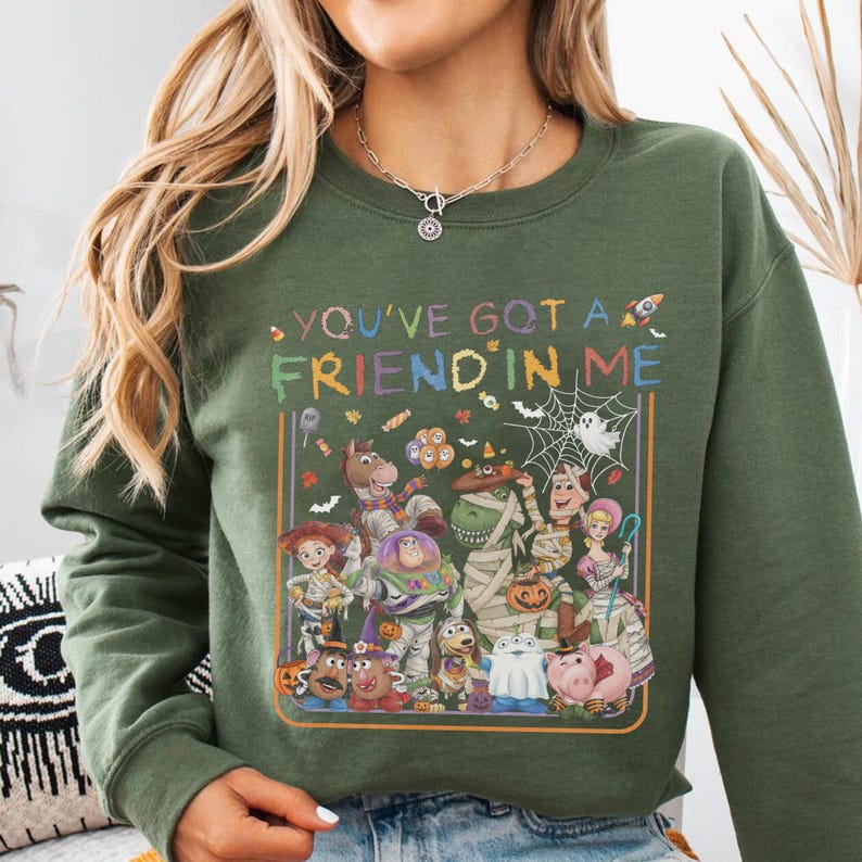 K&ouml;nnte beinhalten: Ein olivgr&uuml;nes Sweatshirt mit Rundhalsausschnitt und einem farbenfrohen Grafikdruck verschiedener Zeichentrickfiguren in Halloween-Kost&uuml;men. Der Text "YOU'VE GOT A FRIEND IN ME" steht &uuml;ber den Figuren, umgeben von kleinen Symbolen wie S&uuml;&szlig;igkeiten, Flederm&auml;usen und einem Spinnennetz.