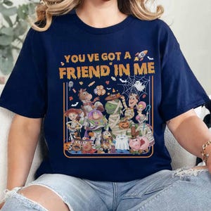 May include: A navy blue t-shirt featuring the orange text "YOU'VE GOT A FRIEND IN ME" above a graphic of various cartoon characters dressed in Halloween costumes, including a mummy, a ghost, and a rocket ship, all framed by an orange border.