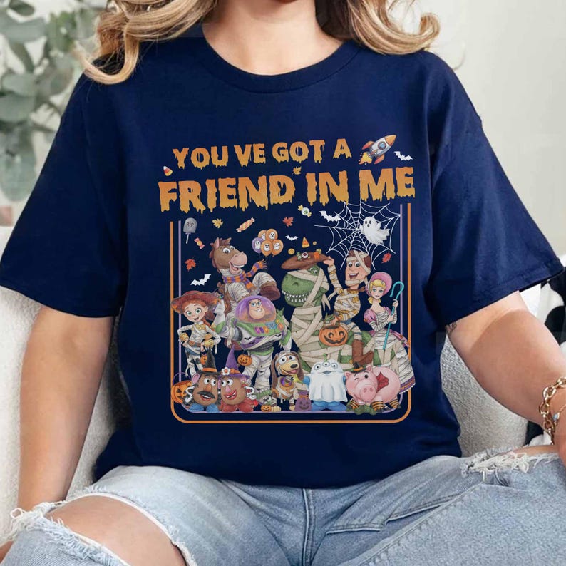 May include: A navy blue t-shirt featuring the orange text "YOU'VE GOT A FRIEND IN ME" above a graphic of various cartoon characters dressed in Halloween costumes, including a mummy, a ghost, and a rocket ship, all framed by an orange border.
