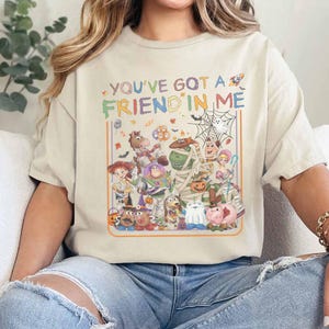 K&ouml;nnte beinhalten: Ein cremefarbenes T-Shirt mit einem Grafikdruck, der den Text "YOU'VE GOT A FRIEND IN ME" &uuml;ber einer Gruppe von Zeichentrickfiguren in Halloween-Kost&uuml;men zeigt, einschlie&szlig;lich eines Spinnennetzes, K&uuml;rbissen und Flederm&auml;usen in einem rechteckigen Rahmen.