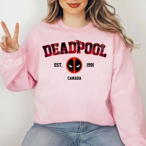 May include: A pale pink sweatshirt with the word "DEADPOOL" in a red and black floral design. Below the word is the text "EST. 1991 CANADA" with a Deadpool logo in the centre. The sweatshirt is a crew neck style.