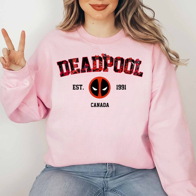 May include: A pale pink sweatshirt with the word "DEADPOOL" in a red and black floral design. Below the word is the text "EST. 1991 CANADA" with a Deadpool logo in the centre. The sweatshirt is a crew neck style.