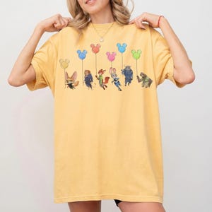 May include: Yellow t-shirt featuring a colorful graphic of cartoon characters holding Mickey Mouse-shaped balloons. The characters are from the movie Zootopia. The shirt has a relaxed fit and short sleeves.