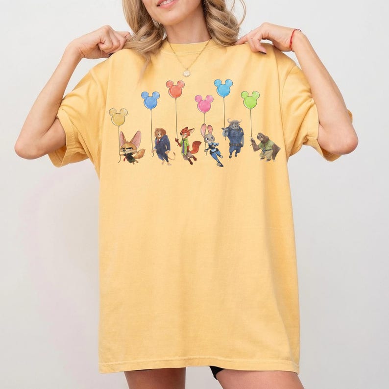 May include: Yellow t-shirt featuring a colorful graphic of cartoon characters holding Mickey Mouse-shaped balloons. The characters are from the movie Zootopia. The shirt has a relaxed fit and short sleeves.