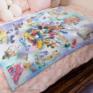 Op de afbeelding: Een kleurrijke Disney-deken met Mickey Mouse, Minnie Mouse, Donald Duck en andere personages. De deken heeft een lichtblauwe en paarse achtergrond met de naam "David" in blauw schrift. Verschillende Disney-personages zijn over de deken verspreid.