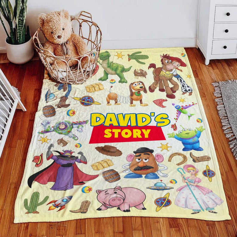 Puede incluir: Una manta rectangular de forro polar amarillo claro con un estampado colorido de varios personajes y objetos de dibujos animados, con el texto "DAVID'S STORY" impreso en letras grandes azules y rojas en el centro de la tela.