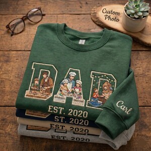 May include: A stack of green, navy, and gray sweatshirts with embroidered designs. The top green sweatshirt features the word "DAD" with a family scene and "EST. 2020". The sleeve is embroidered with the name "Carl".