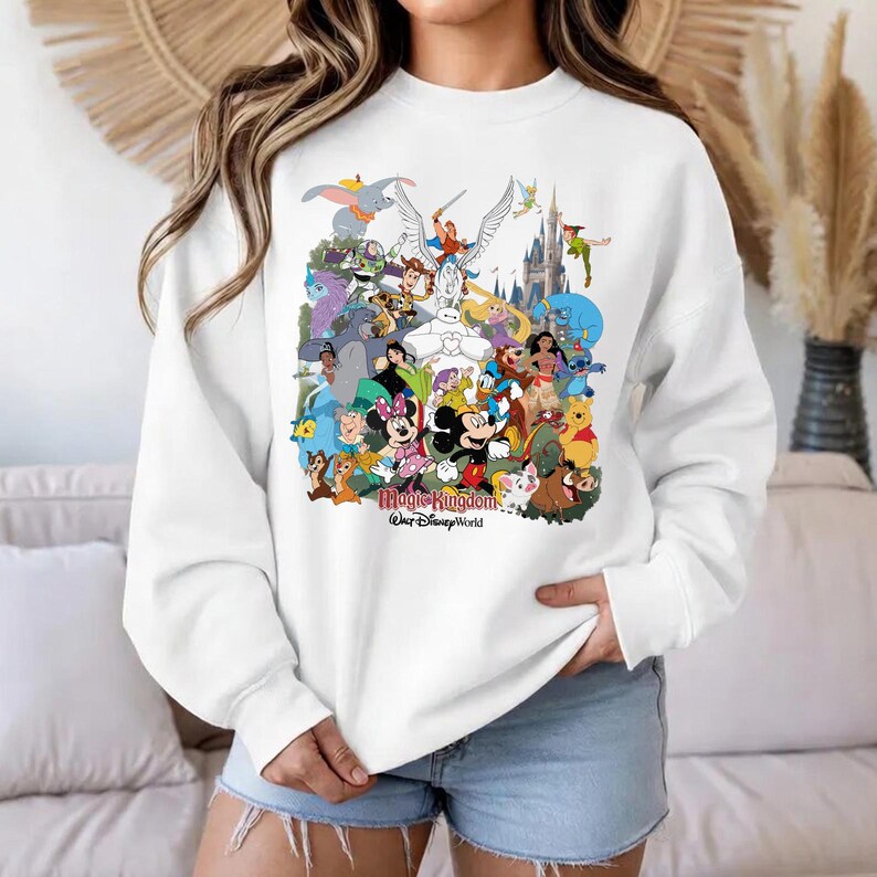Puede incluir: Sudadera blanca con un collage colorido de personajes de Disney, incluyendo Mickey y Minnie Mouse, Dumbo y Peter Pan. El texto "Magic Kingdom, Walt Disney World" est&aacute; impreso en la parte inferior del dise&ntilde;o.