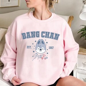 May include: A pale pink sweatshirt featuring the text "BANG CHAN" in blue. Below is a cartoon wolf head, the text "EST. 1997" and "Stray Kids". The sweatshirt has a crew neck design.