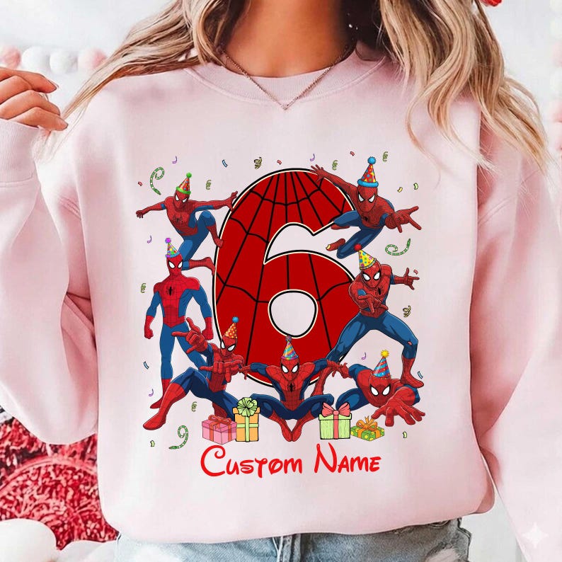 May include: A light pink crewneck sweatshirt featuring a large red number six with a black spiderweb pattern. The design includes multiple illustrations of a red and blue superhero wearing party hats, colourful gift boxes, and the text Custom Name.
