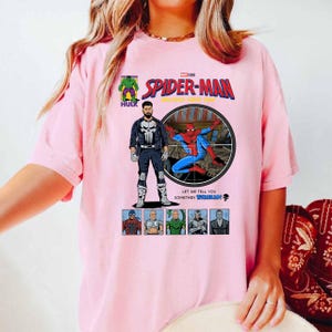 May include: A light pink t-shirt featuring a comic book-style graphic of Spider-Man. The design includes the text "SPIDER-MAN" and images of The Punisher, Hulk, and other characters. The shirt has a crew neck and short sleeves.