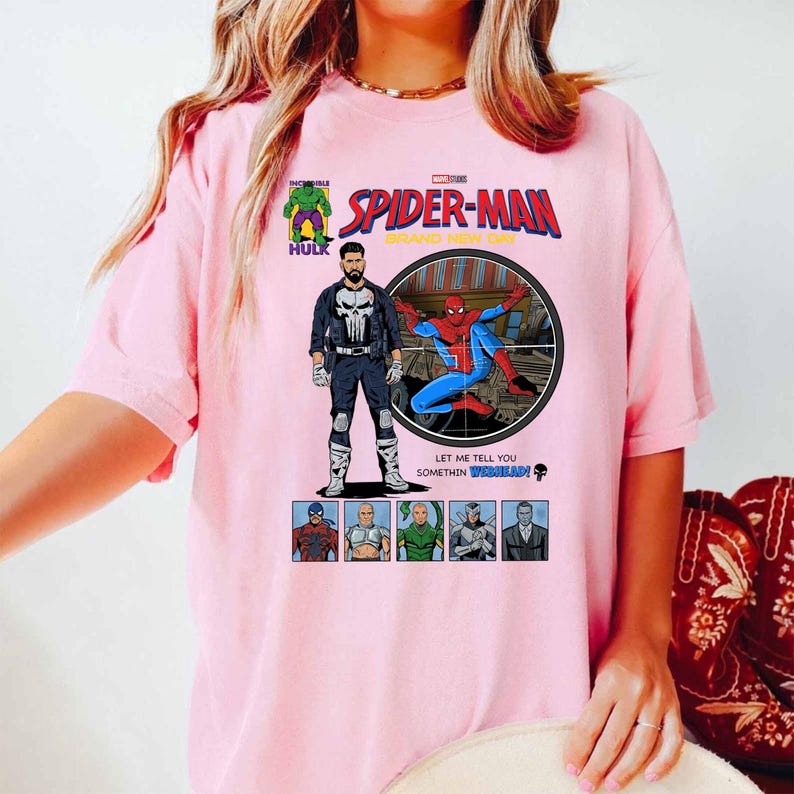 May include: A light pink t-shirt featuring a comic book-style graphic of Spider-Man. The design includes the text "SPIDER-MAN" and images of The Punisher, Hulk, and other characters. The shirt has a crew neck and short sleeves.