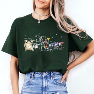 K&ouml;nnte beinhalten: Dunkelgr&uuml;nes T-Shirt mit einer farbenfrohen Grafik von Charakteren aus The Nightmare Before Christmas. Das Design umfasst Jack Skellington, Sally und andere Charaktere, mit Text &uuml;ber und unter den Charakteren.