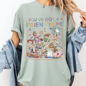 K&ouml;nnte beinhalten: Ein salbeigr&uuml;nes Kurzarm-T-Shirt mit einer bunten Grafik verschiedener Zeichentrickfiguren in Halloween-Kost&uuml;men. Der Text "YOU'VE GOT A FRIEND IN ME" steht &uuml;ber den Figuren, umgeben von kleinen Symbolen wie S&uuml;&szlig;igkeiten und einem Spinnennetz.
