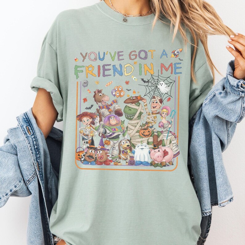 K&ouml;nnte beinhalten: Ein salbeigr&uuml;nes Kurzarm-T-Shirt mit einer bunten Grafik verschiedener Zeichentrickfiguren in Halloween-Kost&uuml;men. Der Text "YOU'VE GOT A FRIEND IN ME" steht &uuml;ber den Figuren, umgeben von kleinen Symbolen wie S&uuml;&szlig;igkeiten und einem Spinnennetz.
