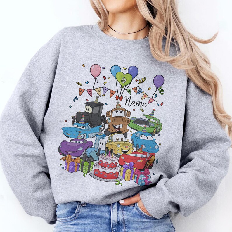 Peut inclure: Un sweat-shirt gris clair avec un motif color&eacute; de voitures de dessins anim&eacute;s, de ballons, de confettis et d'un g&acirc;teau d'anniversaire. Le mot "Name" est &eacute;crit en cursive. Le motif comprend divers personnages de voitures et des bo&icirc;tes cadeaux.