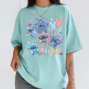 Może przedstawiać: Jasnoniebieska koszulka z grafiką postaci Stitch z filmu Lilo & Stitch. Stitch jest przedstawiony w ramkach w kształcie gwiazd i trzymający balony. Słowo "magical" jest napisane kolorowymi literami poniżej.