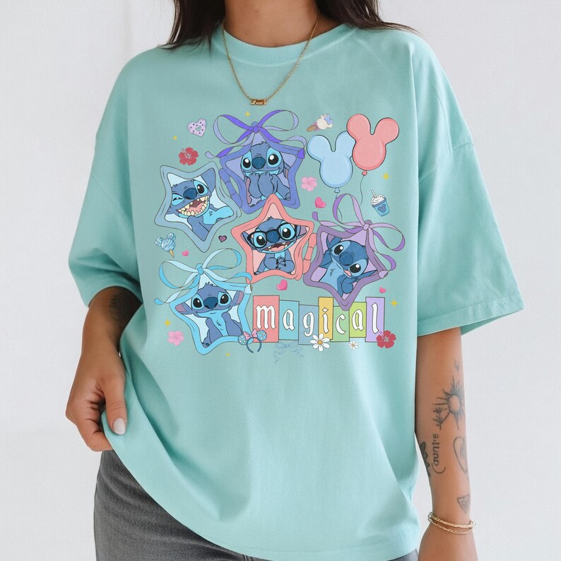 Może przedstawiać: Jasnoniebieska koszulka z grafiką postaci Stitch z filmu Lilo & Stitch. Stitch jest przedstawiony w ramkach w kształcie gwiazd i trzymający balony. Słowo "magical" jest napisane kolorowymi literami poniżej.
