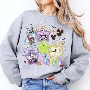 May include: A light grey sweatshirt with a colourful graphic design. The design features star-shaped frames with characters, bows, bats, and the word "magical". The sweatshirt is made of a soft material.