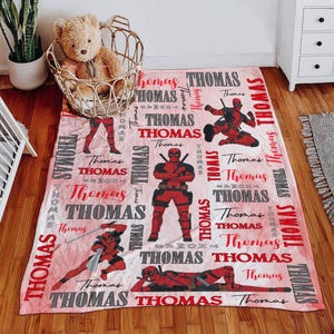 May include: A pink blanket featuring multiple images of a red and black superhero in various action poses. The name "Thomas" is repeated throughout the design in red and gray lettering.