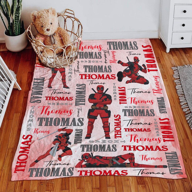 May include: A pink blanket featuring multiple images of a red and black superhero in various action poses. The name "Thomas" is repeated throughout the design in red and gray lettering.