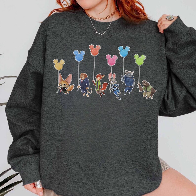 May include: Dark gray sweatshirt featuring a colorful graphic of cartoon characters holding Mickey Mouse-shaped balloons. The characters include a fox, a lion, a rabbit, and others. The balloons are yellow, blue, orange, pink, and green.