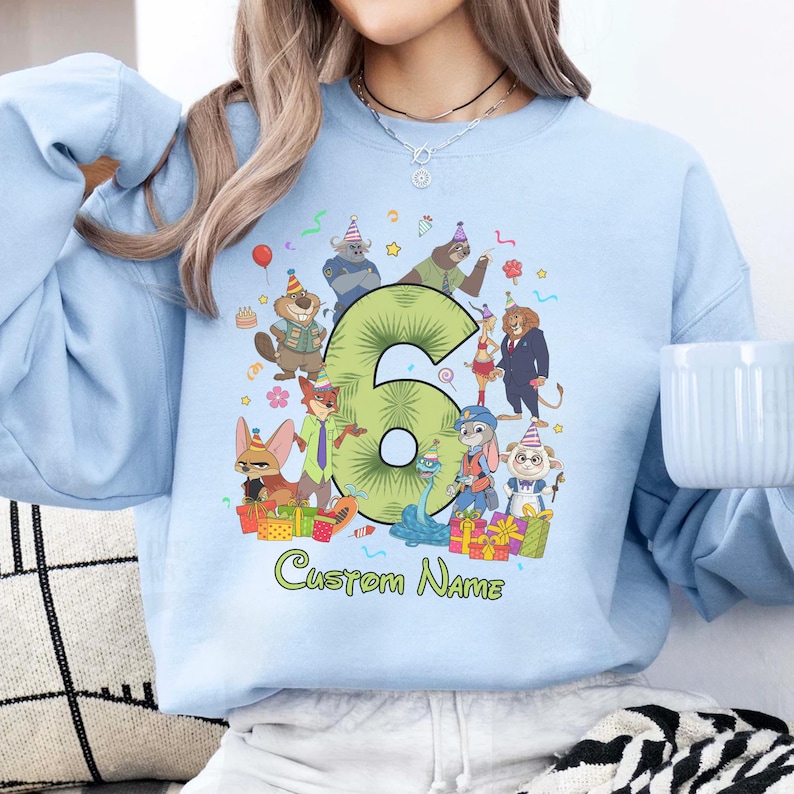 May include: A light blue crewneck sweatshirt featuring a large green number six surrounded by various cartoon animal characters wearing party hats, colorful gift boxes, and confetti. The text Custom Name is printed in green below the graphic.