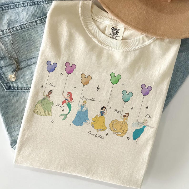 May include: Cream-colored t-shirt featuring Disney princesses holding Mickey Mouse-shaped balloons. The princesses include Tiana, Ariel, Cinderella, Snow White, Belle, and Elsa. The shirt has the Comfort Colors brand tag.