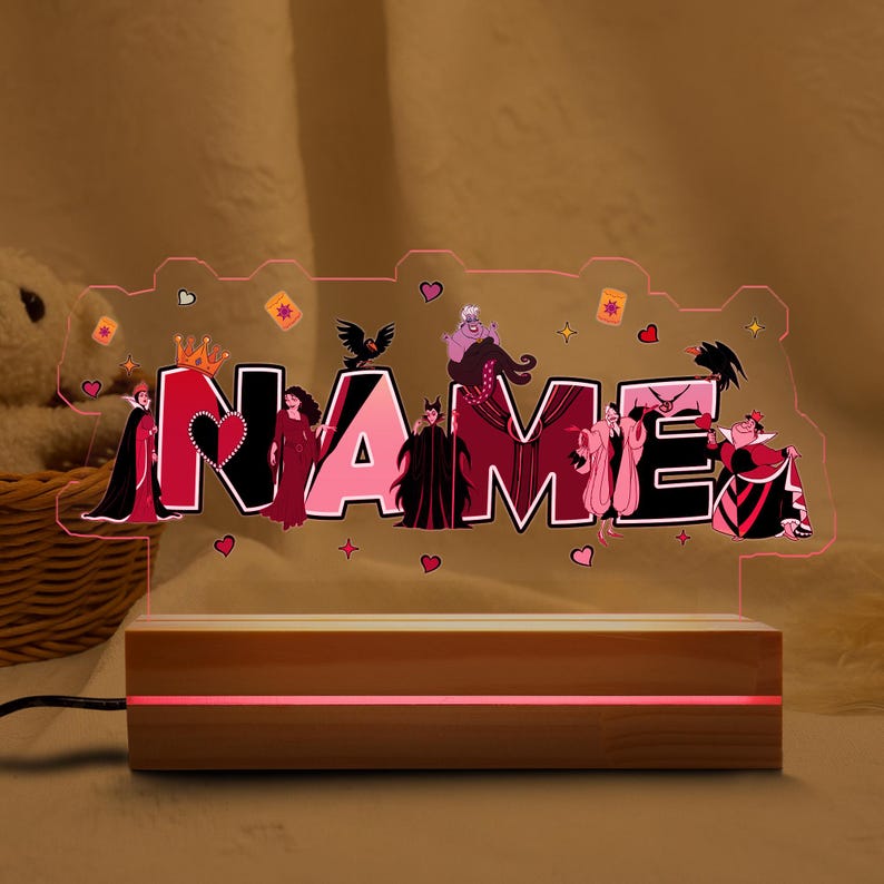May include: A personalized night light with the name "NAME" illuminated in pink. The design features cartoon villains and decorative hearts, stars, and crowns. The light sits on a wooden base with a red glow.