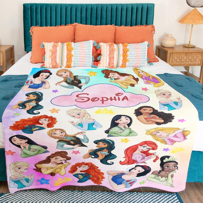 May include: A personalized blanket featuring various cartoon princesses in a colorful design. The blanket has a cloud with the name "Sophia" in pink. The princesses are in different poses and outfits, with stars and crowns.