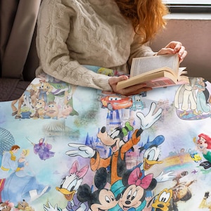 Op de afbeelding: Een kleurrijke deken met Disney-personages zoals Mickey Mouse, Donald Duck en Assepoester. De deken heeft een aquarelontwerp met afbeeldingen van verschillende Disney-films en -personages. Een persoon houdt de deken vast terwijl hij een boek leest.