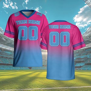Puede incluir: Dos camisetas de f&uacute;tbol rosas y azules. La camiseta delantera tiene el texto "TEAM NAME" encima del n&uacute;mero "00". La camiseta trasera tiene el texto "YOUR NAME" encima del n&uacute;mero "00". Ambas camisetas tienen detalles azules y cuello en V.
