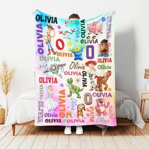 Op de afbeelding: Een zachte fleece deken met een kleurrijke tie-dye achtergrond en illustraties van diverse Toy Story-personages zoals Woody, Buzz Lightyear, Jessie, Rex en Hamm. De naam Olivia staat herhaaldelijk in verschillende lettertypes gedrukt.