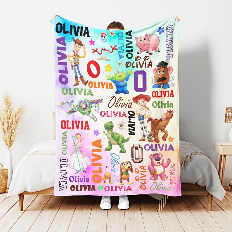 Op de afbeelding: Een zachte fleece deken met een kleurrijke tie-dye achtergrond en illustraties van diverse Toy Story-personages zoals Woody, Buzz Lightyear, Jessie, Rex en Hamm. De naam Olivia staat herhaaldelijk in verschillende lettertypes gedrukt.