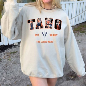 May include: Beige sweatshirt with the word "TANO" in orange and black block letters, featuring character illustrations. Below, the text reads "EST. 36 BBY THE CLONE WARS" in orange. The sweatshirt has long sleeves.