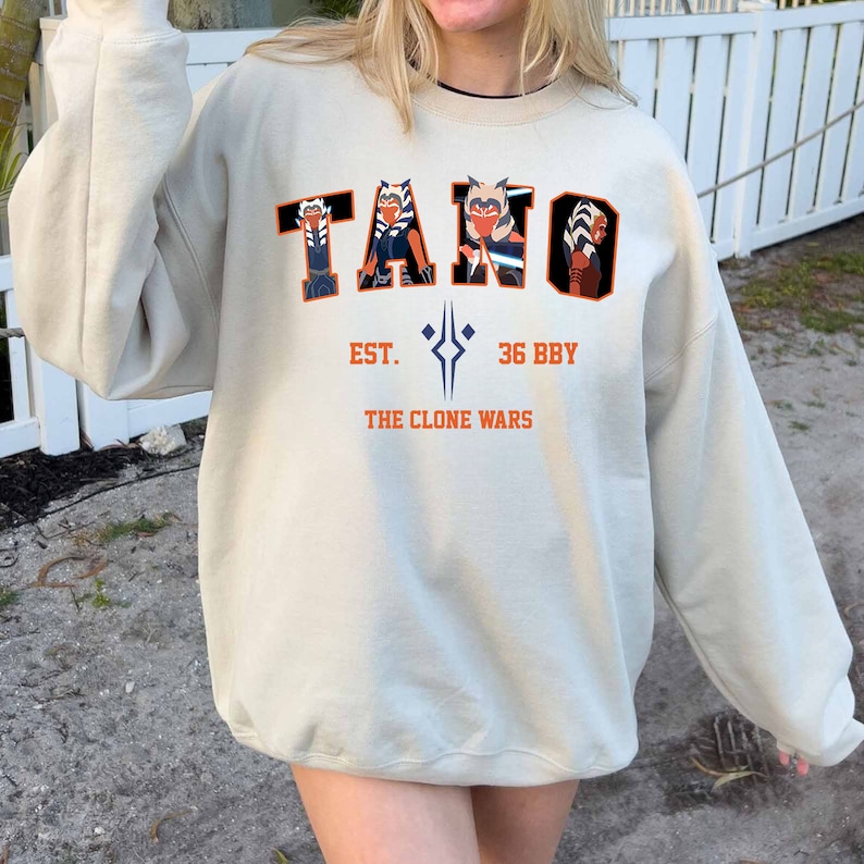 May include: Beige sweatshirt with the word "TANO" in orange and black block letters, featuring character illustrations. Below, the text reads "EST. 36 BBY THE CLONE WARS" in orange. The sweatshirt has long sleeves.