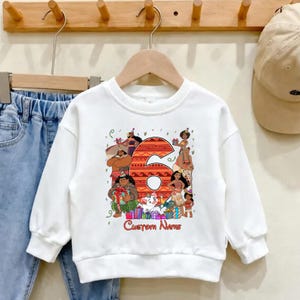 May include: A white long-sleeve sweatshirt featuring a large orange patterned number six surrounded by various animated characters from the movie Moana, including Maui and Moana, with colorful gift boxes and the text Custom Name printed below.