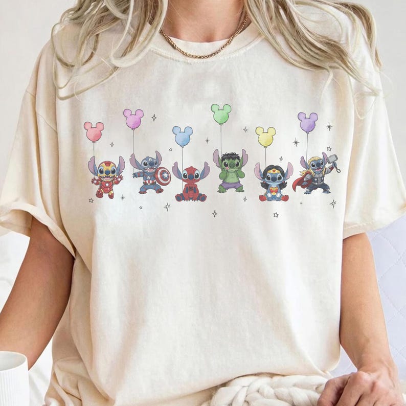 May include: Cream-colored t-shirt featuring a colorful graphic of cartoon characters dressed as superheroes, each holding a Mickey Mouse-shaped balloon. The characters include Iron Man, Captain America, Deadpool, Hulk, Wonder Woman, and Thor.