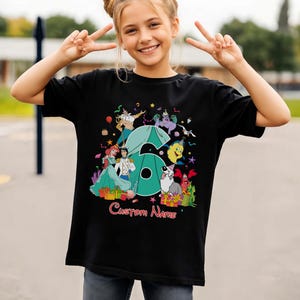 K&ouml;nnte beinhalten: Ein schwarzes Kurzarm-T-Shirt mit einer gro&szlig;en t&uuml;rkisfarbenen Zahl Sechs, umgeben von bunten Zeichentrickfiguren aus Arielle, die Meerjungfrau, darunter Arielle, Prinz Erik und Sebastian, mit dem Text Custom Name in roter Schrift unten.
