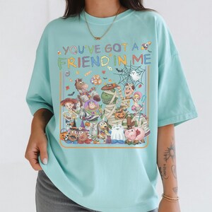 K&ouml;nnte beinhalten: Ein helles t&uuml;rkisfarbenes T-Shirt mit einem farbenfrohen Grafikdruck verschiedener Zeichentrickfiguren in Halloween-Kost&uuml;men. Der Text "YOU'VE GOT A FRIEND IN ME" ist &uuml;ber den Figuren aufgedruckt, begleitet von kleinen Symbolen einer Rakete, S&uuml;&szlig;igkeiten und einem Spinnennetz.
