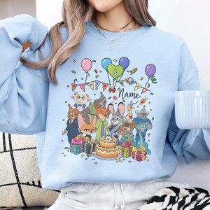 Puede incluir: Sudadera azul claro con una ilustraci&oacute;n de dibujos animados de personajes, una tarta de cumplea&ntilde;os, globos y confeti. La palabra "Name" est&aacute; escrita en cursiva. La sudadera es de estilo casual.