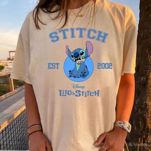 May include: A beige t-shirt featuring a blue graphic of Stitch from Disney's Lilo & Stitch. The design includes the word "STITCH" above the character, with "EST 2002" and the Disney logo below. The shirt has short sleeves.