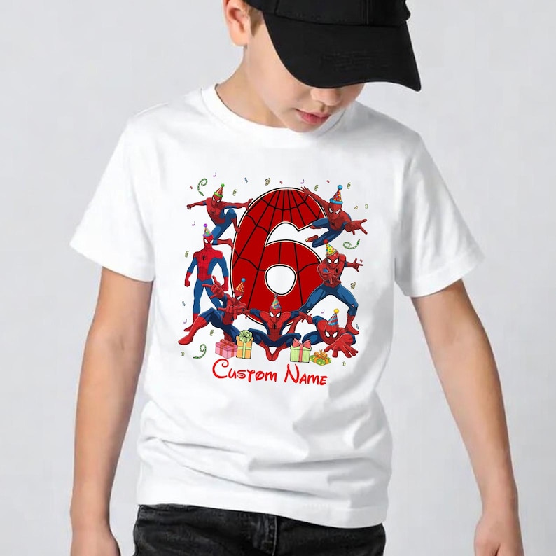 May include: A white t-shirt featuring a large red number six with a spiderweb pattern, surrounded by multiple illustrations of Spider-Man wearing colourful party hats. Small gift boxes and confetti are present, with the text Custom Name below.