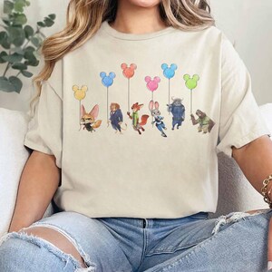 May include: Beige t-shirt featuring a watercolor illustration of characters from a popular animated film, each holding a Mickey Mouse-shaped balloon in various colors. The design includes a fox, a lion, a fox, a rabbit, a buffalo, and a sloth.