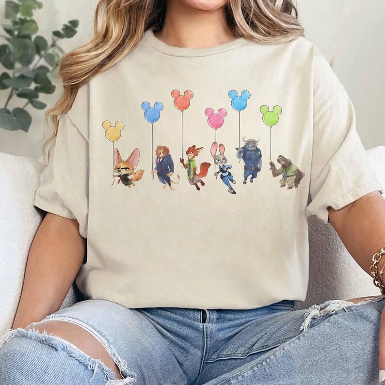May include: Beige t-shirt featuring a watercolor illustration of characters from a popular animated film, each holding a Mickey Mouse-shaped balloon in various colors. The design includes a fox, a lion, a fox, a rabbit, a buffalo, and a sloth.