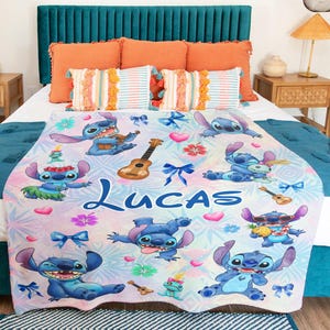 Op de afbeelding: Een witte deken met een herhalend patroon van het personage Stitch uit Lilo & Stitch, ukeleles, strikken en bloemen. De naam "Lucas" is in blauwe letters gedrukt. De deken ligt op een bed met oranje en turquoise kussens.