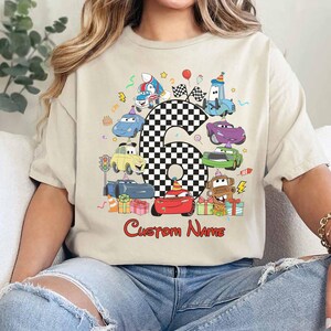 May include: Beige t-shirt with a cartoon car design, featuring the number 6 in a chequered pattern. The shirt has the text "Custom Name" in red. The design includes various car characters and birthday elements, ideal for a sixth birthday celebration.