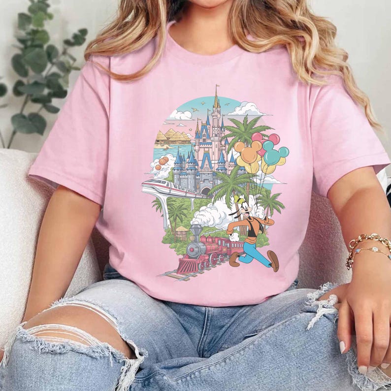 以下が含まれることがあります： テーマパークのカラフルなイラストが描かれた、明るいピンク色のTシャツ。デザインには、お城、電車、ヤシの木、風船、そして走っている漫画のキャラクターが含まれています。このシャツは柔らかい素材で作られています。