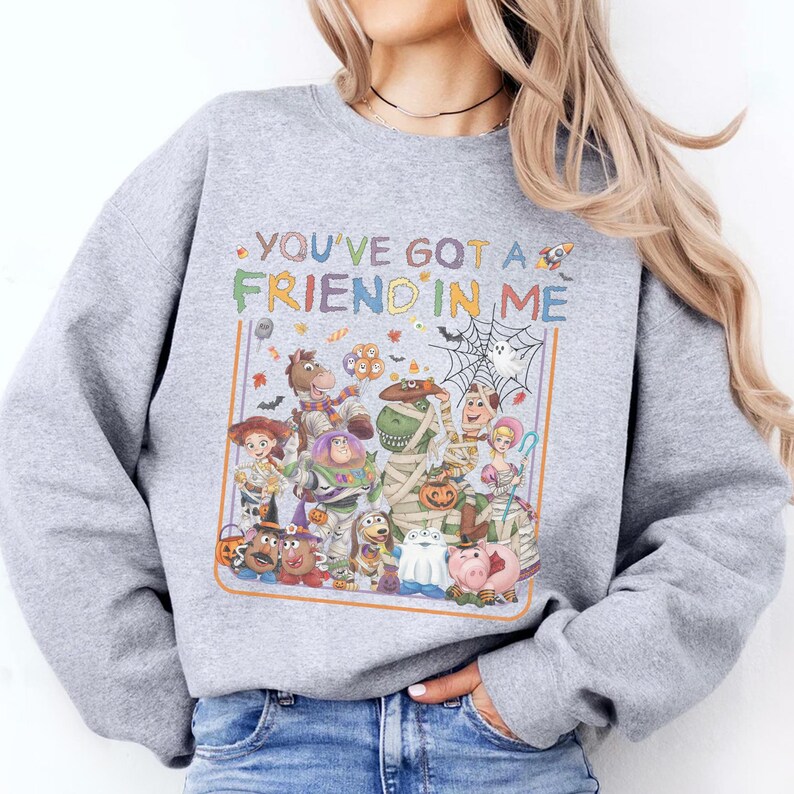 K&ouml;nnte beinhalten: Ein graumeliertes Sweatshirt mit Rundhalsausschnitt und einem farbenfrohen Grafikdruck verschiedener Zeichentrickfiguren in Halloween-Kost&uuml;men. Der Text "YOU'VE GOT A FRIEND IN ME" steht &uuml;ber den Figuren, erg&auml;nzt durch kleine Symbole von S&uuml;&szlig;igkeiten, Flederm&auml;usen und einem Spinnennetz.